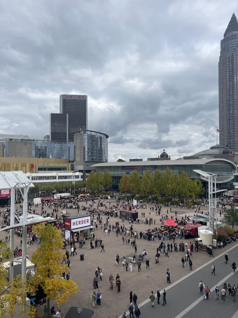 Blick auf den belebten Innenhof der Frankfurter Buchmesse mit Besuchern, Messehallen und Hochhäusern unter bewölktem Himmel.